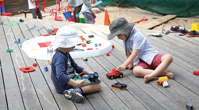 Two children with hats on playing outdoors with a small red truck