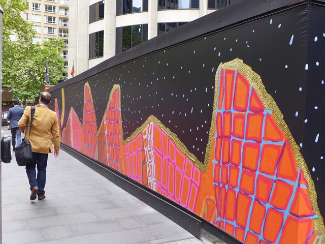 Colourful images in front of a building site on the right, and a footpath with a person walking on it on the left.