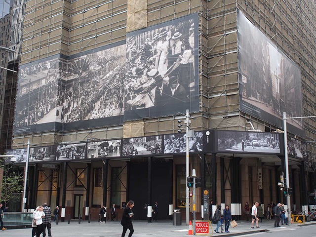 People on a street, a large building in the distance with boards on it.