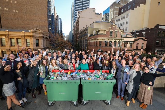 Crowd of happy people in a sunny day gathered around two commercial bins full of plastic celebrating with arms and fists raised in the air.