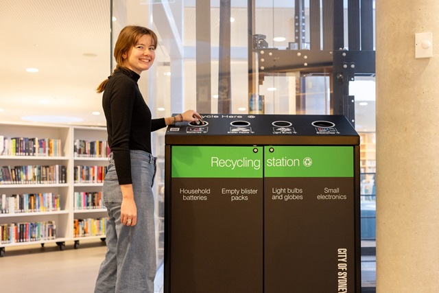 A smiling woman stands by a recycling station in a library, placing an item into one of the bins labelled for different types of recyclable materials.