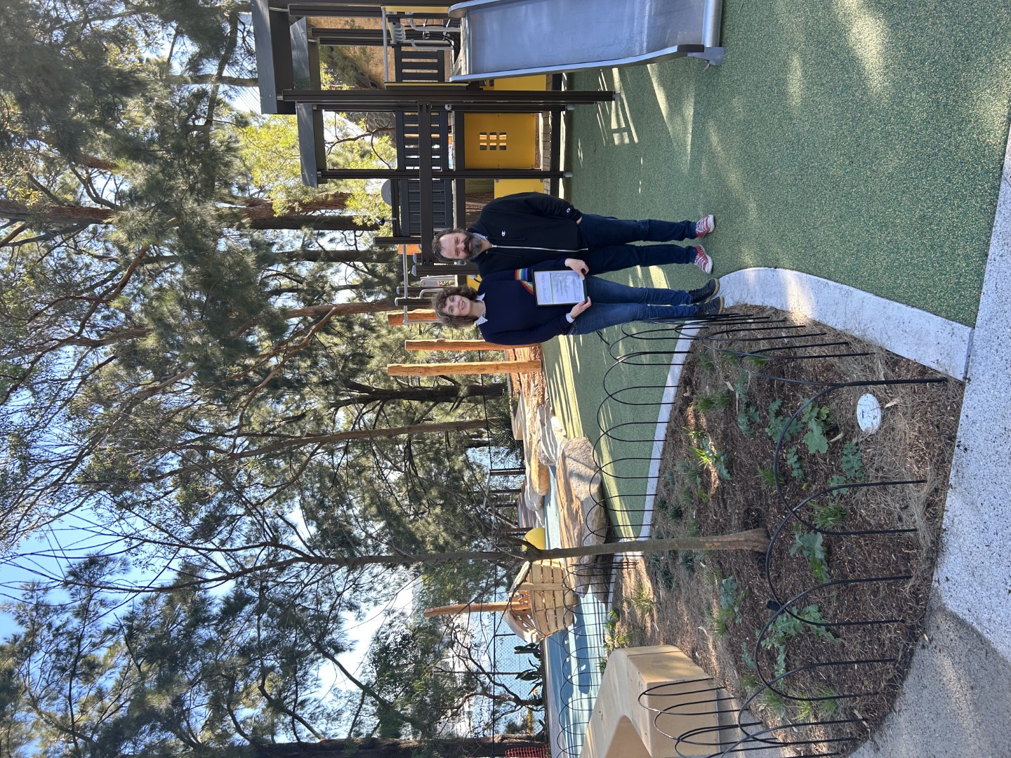 Two people standing in front of a playground next to a tree