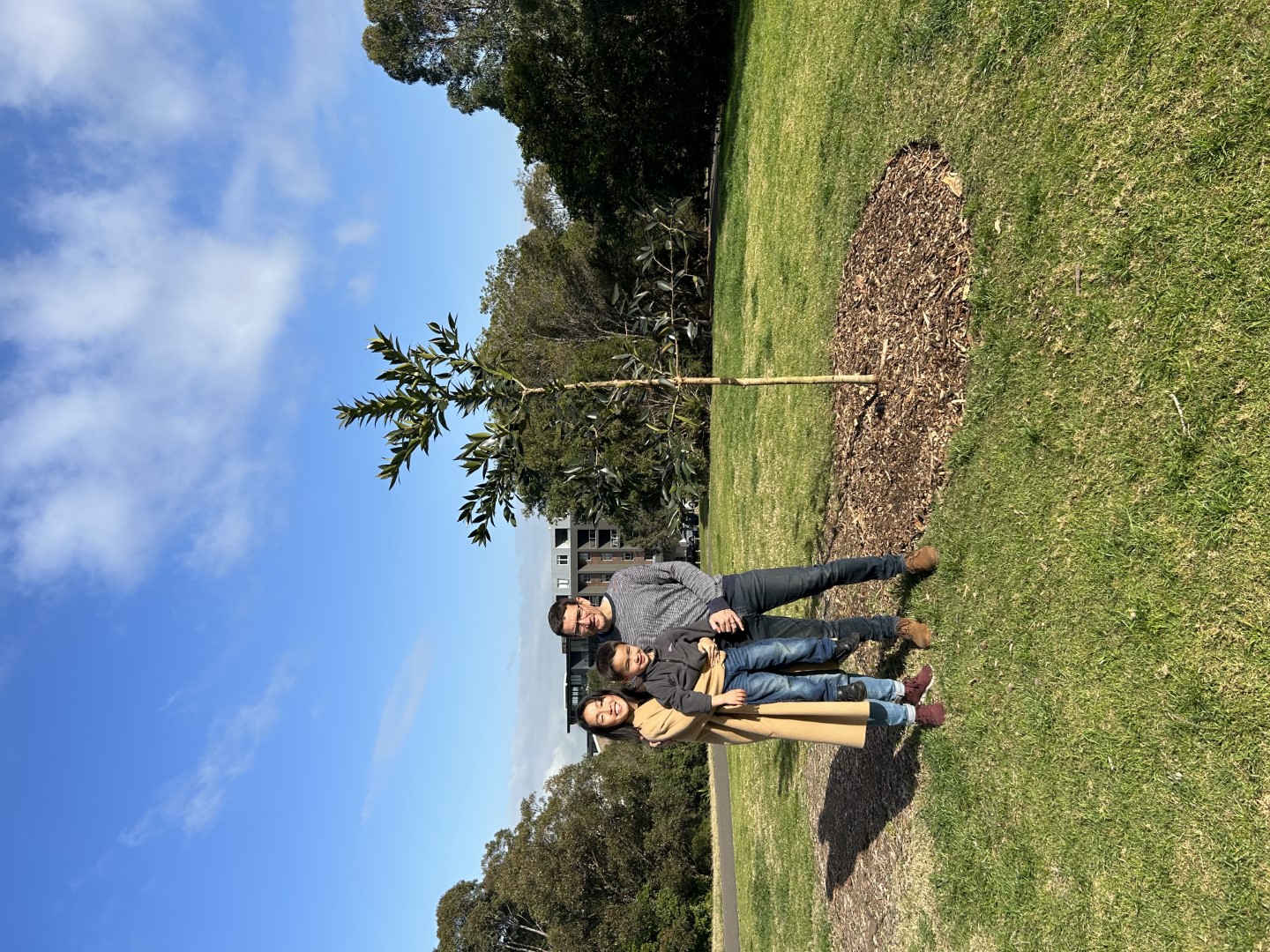 Two adults holding a small child standing next to a tree in a grassy area.