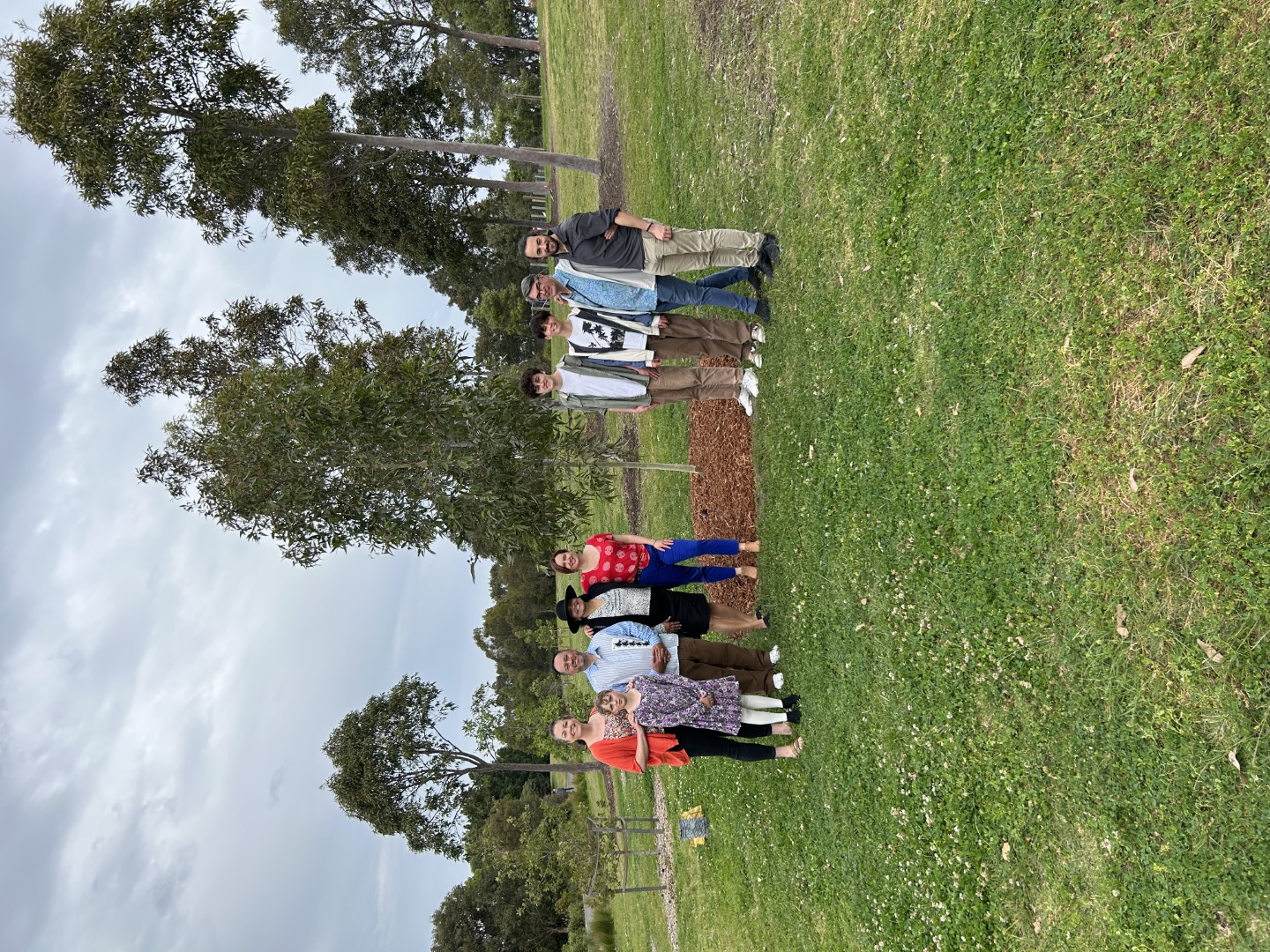 A group of people standing in a semi circle in front of a tree in a park.