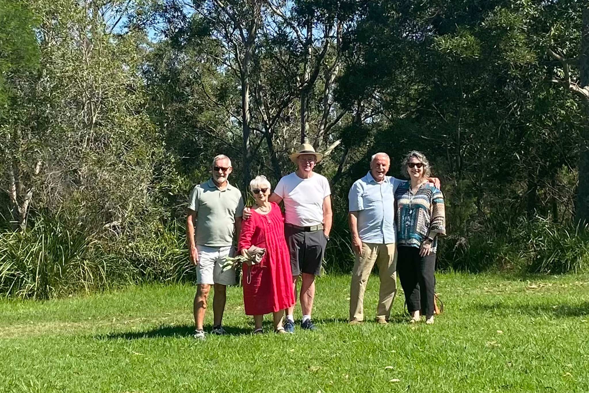 Five adults stand together on grass in a sunny park with trees in the background.