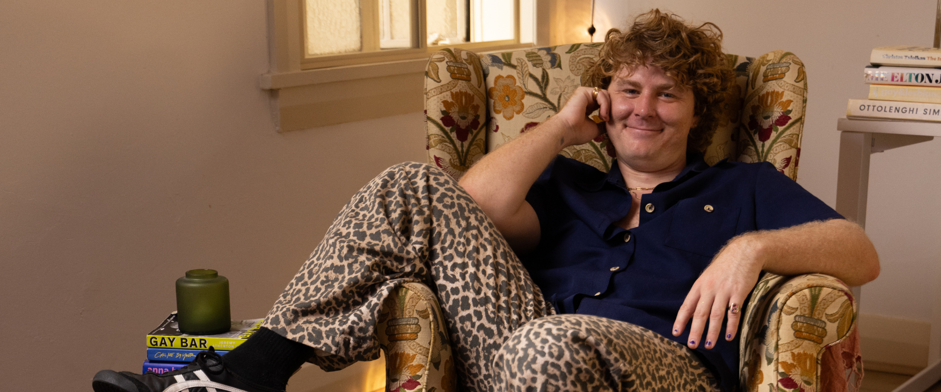 Person with curly hair sits in a patterned armchair, smiling and holding a phone to their ear. They wear a navy shirt and leopard print pants. Books and a candle are stacked nearby.