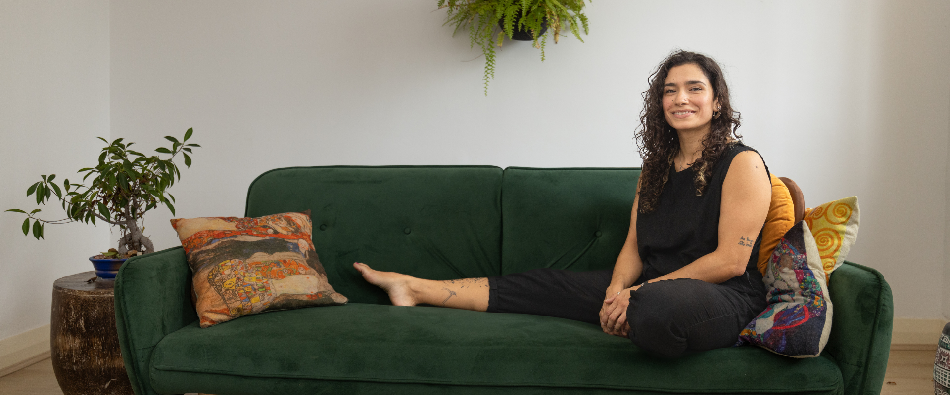 A person with long curly hair sits on a green sofa with one leg extended, surrounded by colorful pillows, a potted plant, and a hanging fern in a well-lit room.