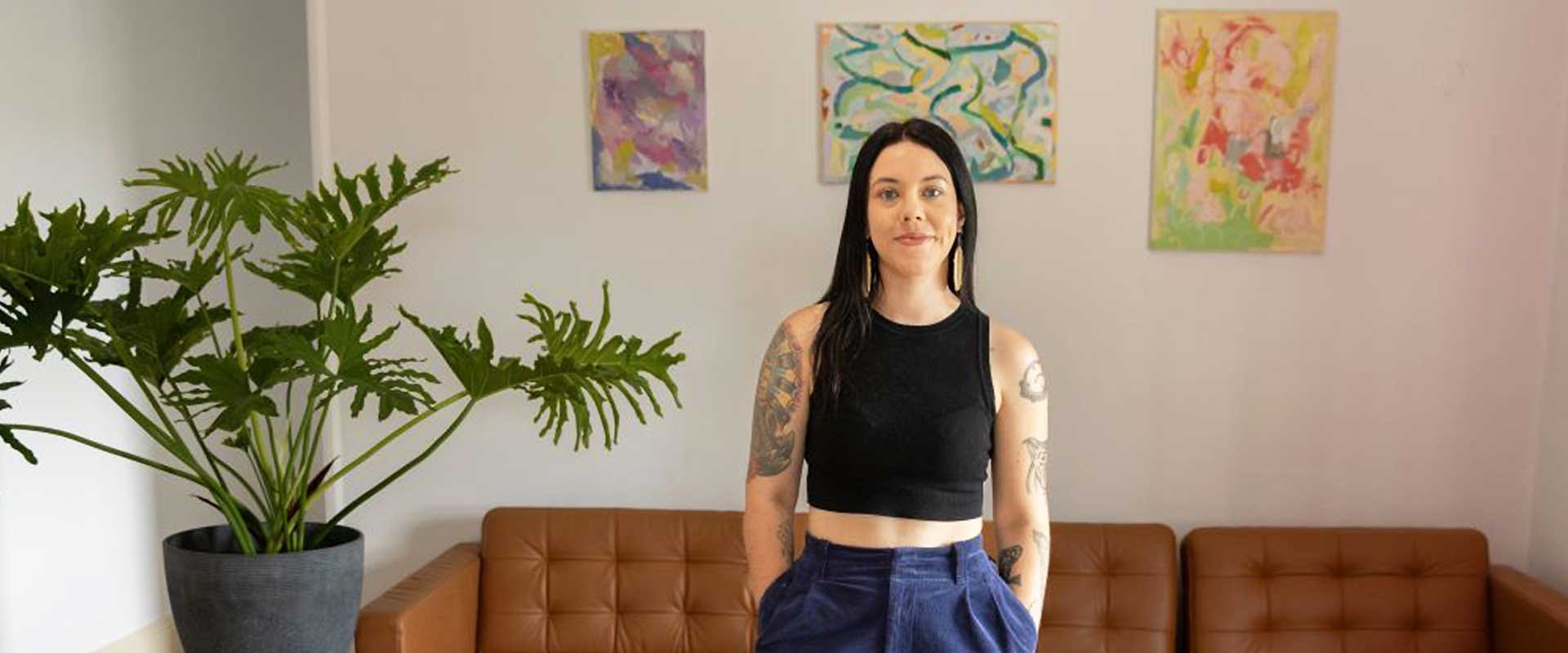 A woman in a black crop top and blue pants stands in a living room with a brown sofa, abstract art on the wall.