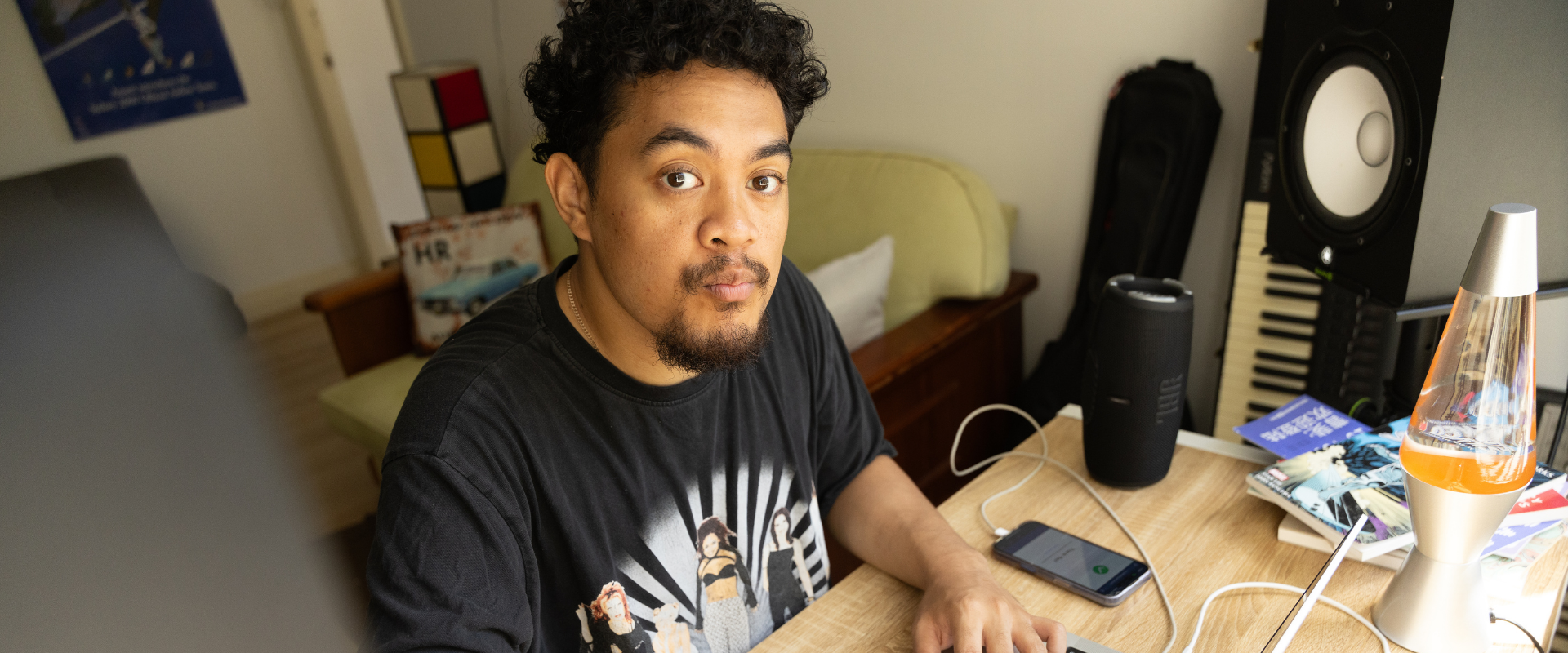 A man with curly hair sits at a desk with a keyboard, speaker, books, smartphone, and a lava lamp, looking directly at the camera.